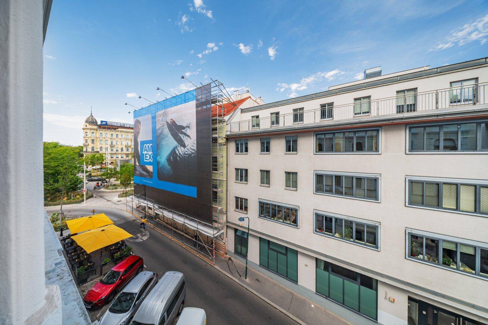 Blick von einem Balkon auf eine belebte Stadtstraße mit Gebäuden und einem großen Werbebanner.