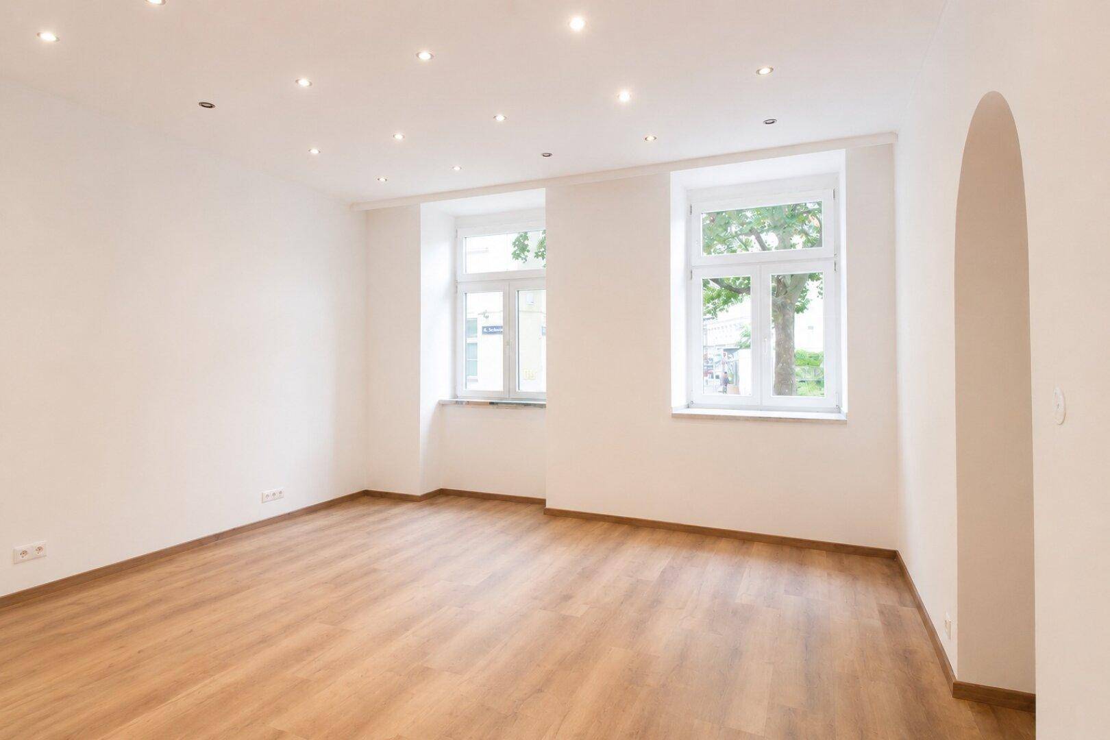 Geräumiger Wohnbereich mit hellem Holzboden, weißen Wänden und Blick ins Grüne durch die Fenster.
