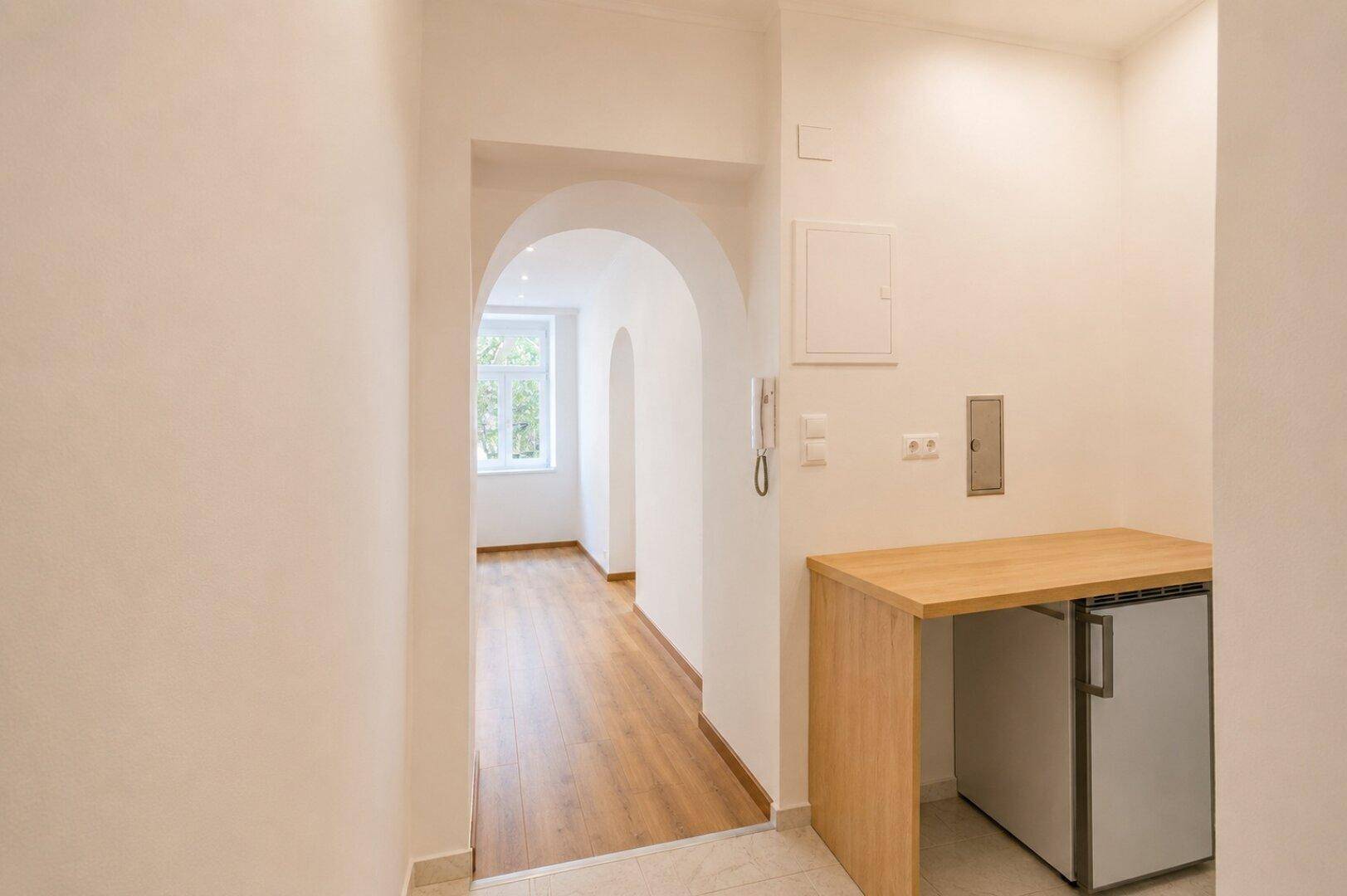 Eingangsbereich mit kleiner Küchenzeile, Kühlschrank und Holzarbeitsplatte, verbunden mit einem Wohnraum.