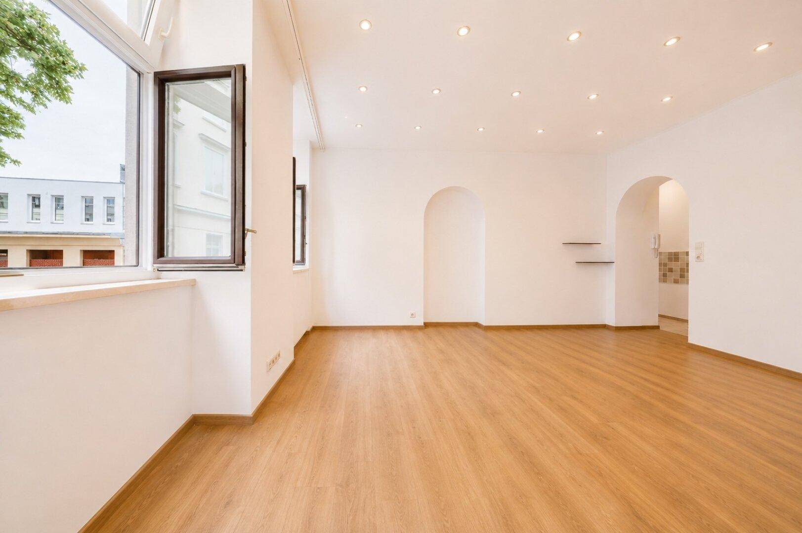 Heller Wohnraum mit Holzboden, weißen Wänden, mehreren Fenstern und Zugang zu einem weiteren Bereich.