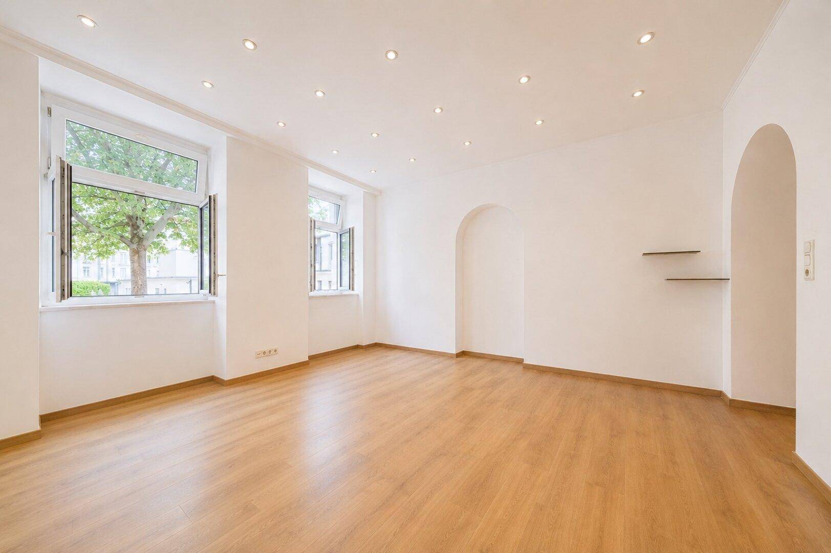 Heller, leerer Raum mit Holzboden, weißen Wänden, zwei Fenstern und Deckenstrahlern.