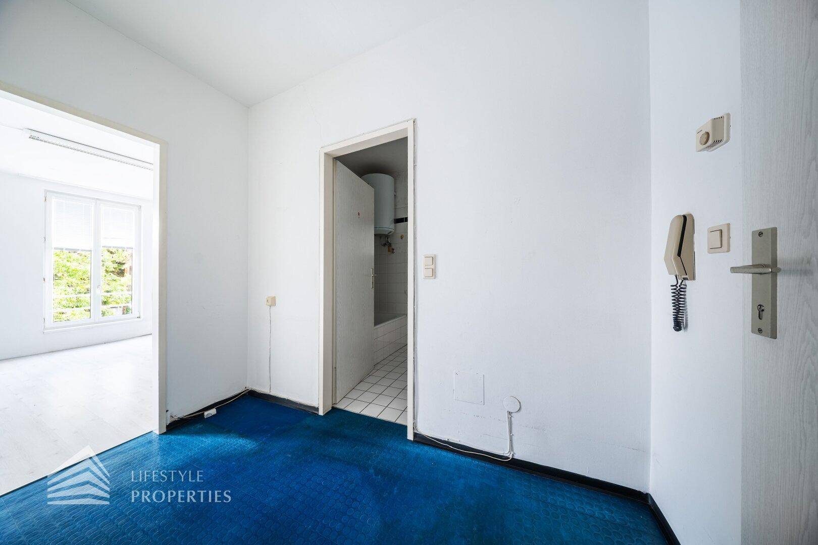 Heller Flur mit blauem Teppichboden und Gegensprechanlage, führt zu weiteren Zimmern.