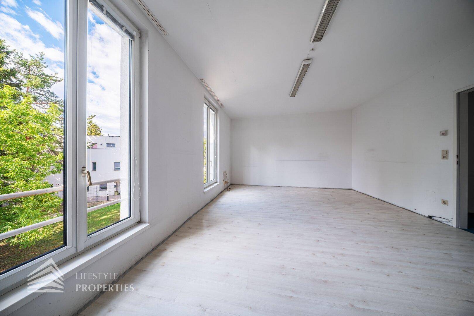 Geräumiges Zimmer mit großen Fenstern, die einen Blick ins Grüne und auf umliegende Gebäude bieten.