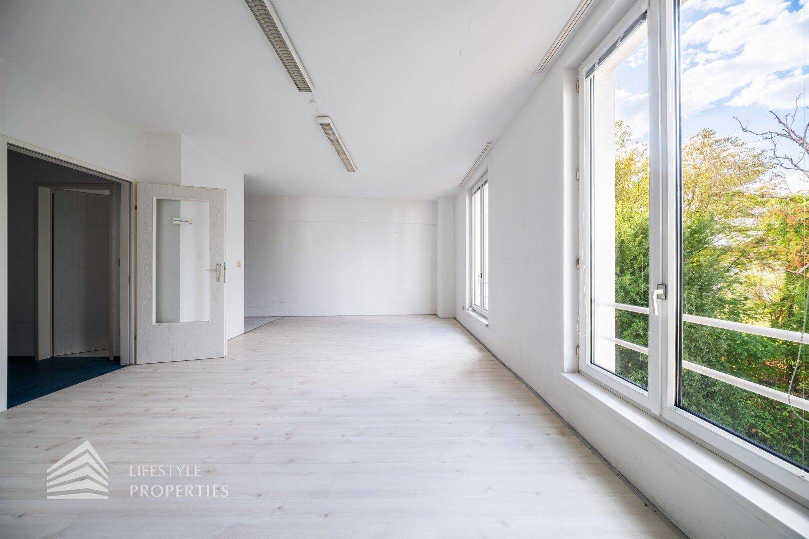 Großes, helles Zimmer mit mehreren Fenstern und Zugang zu einem weiteren Raum.