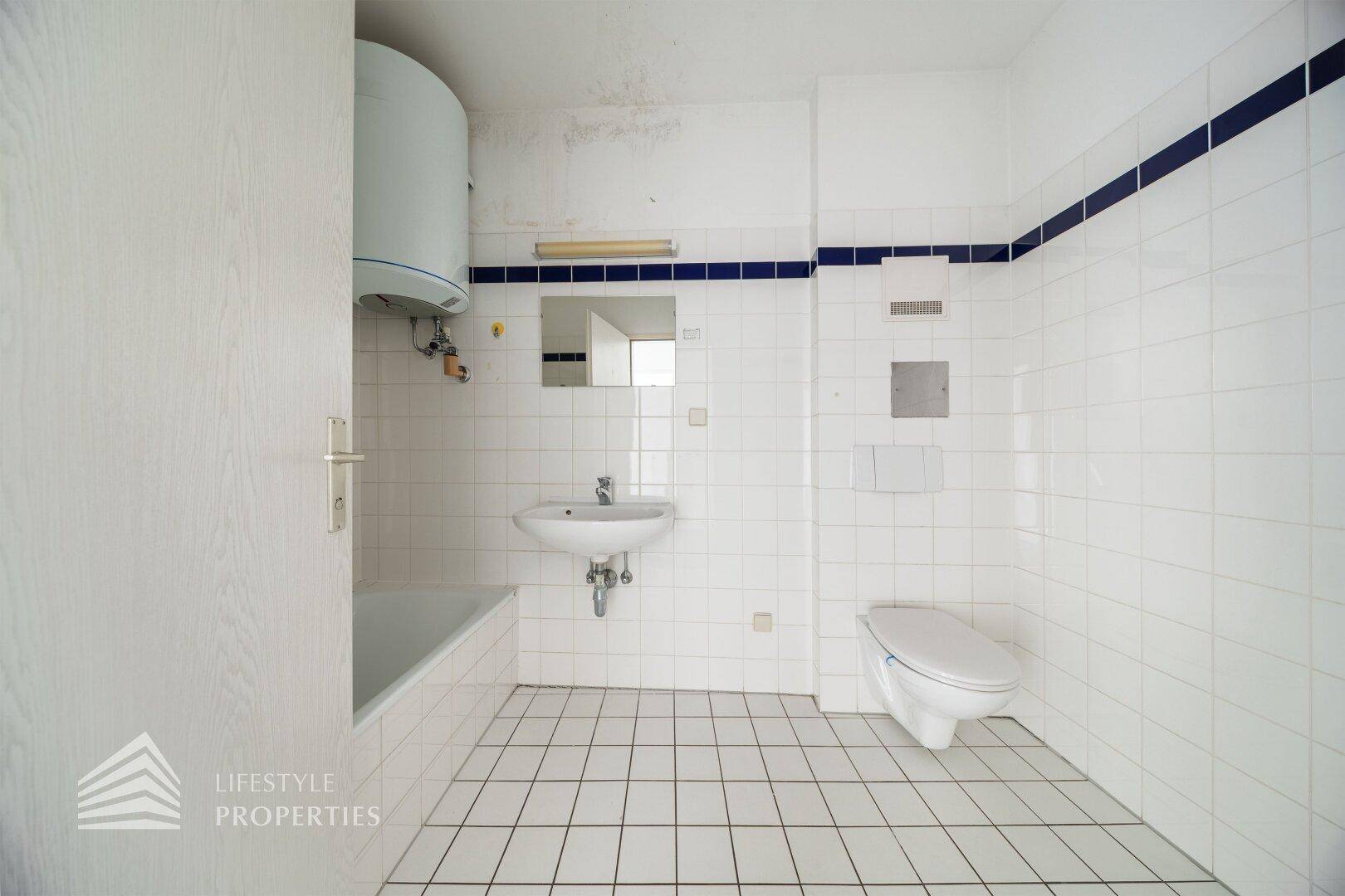 Badezimmer mit WC, Waschbecken und Badewanne, komplett gefliest in Weiß mit blauer Bordüre.