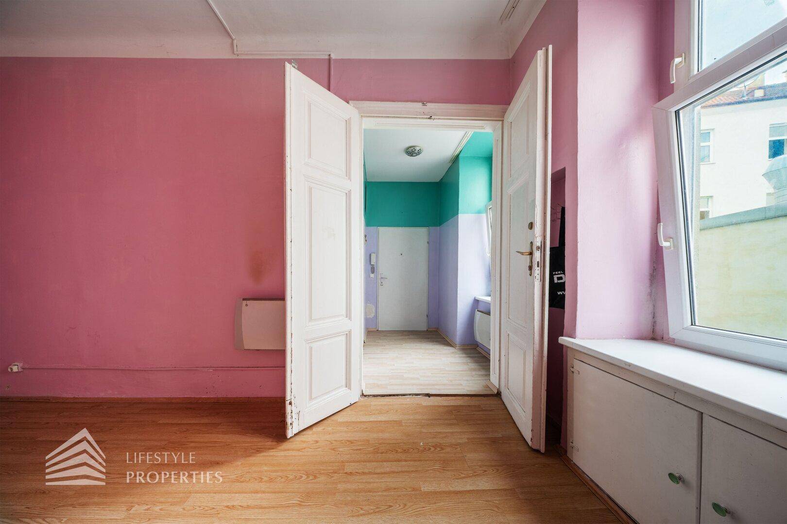 Geräumiges Zimmer mit rosa Wänden, Holzboden und einer offenen Tür zu einem weiteren Raum.