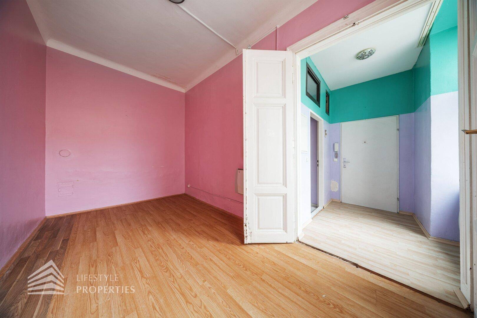 Helles Zimmer mit rosa Wänden, Holzboden und einer offenen Tür, die den Blick in den Flur freigibt.