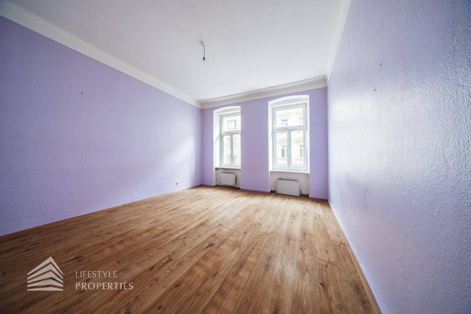 Leeres Zimmer mit lila Wänden, Holzboden und zwei hohen Fenstern, die viel Tageslicht bieten.