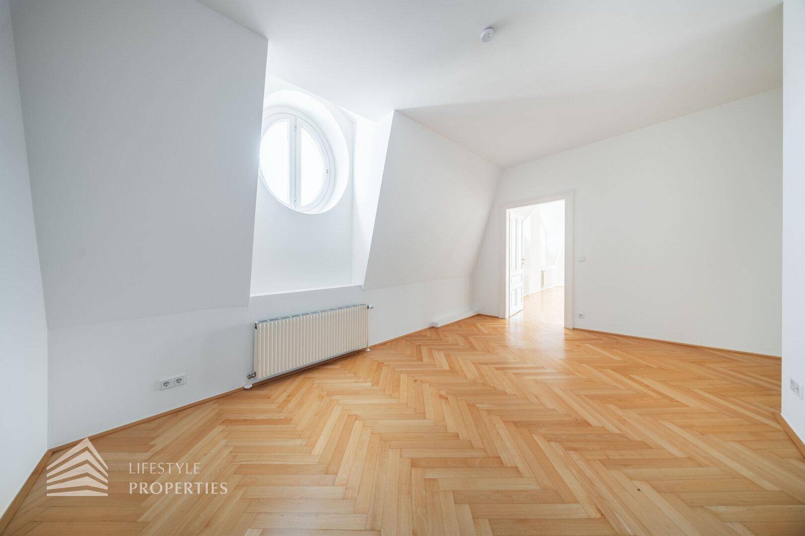 Dachgeschosszimmer mit Fischgrätparkett, weiß gestrichenen Wänden und ovalem Fenster.