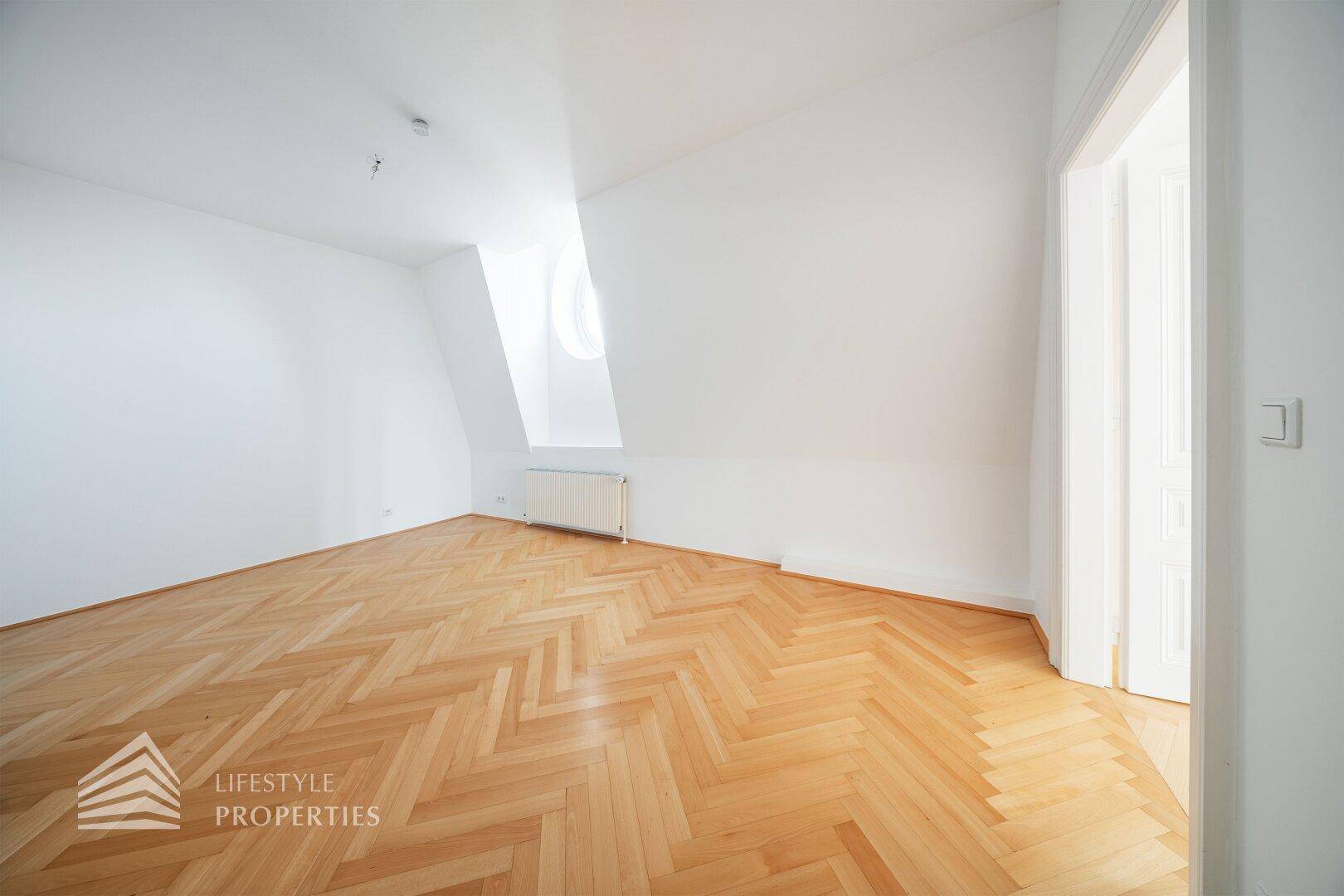 Leeres Dachgeschosszimmer mit Fischgrätparkett und schräger Wand mit Fenster.