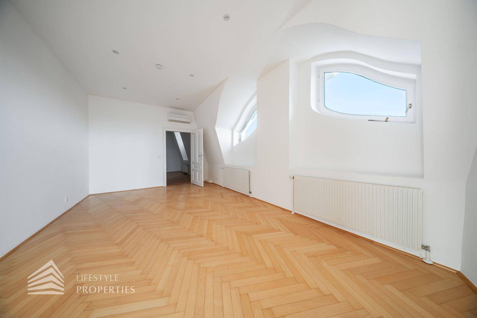 Dachgeschosszimmer mit Fischgrätparkett, weiß gestrichenen Wänden und einem geschwungenen Fenster.