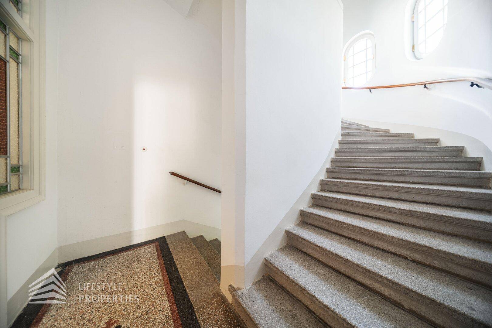 Helles Treppenhaus mit Steinstufen und Buntglasfenstern, das zu den oberen Etagen führt.