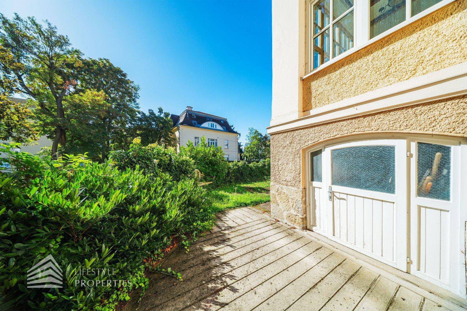 Seitlicher Zugang zur Villa mit Garagentor und einem kleinen Gartenbereich im Sonnenschein.