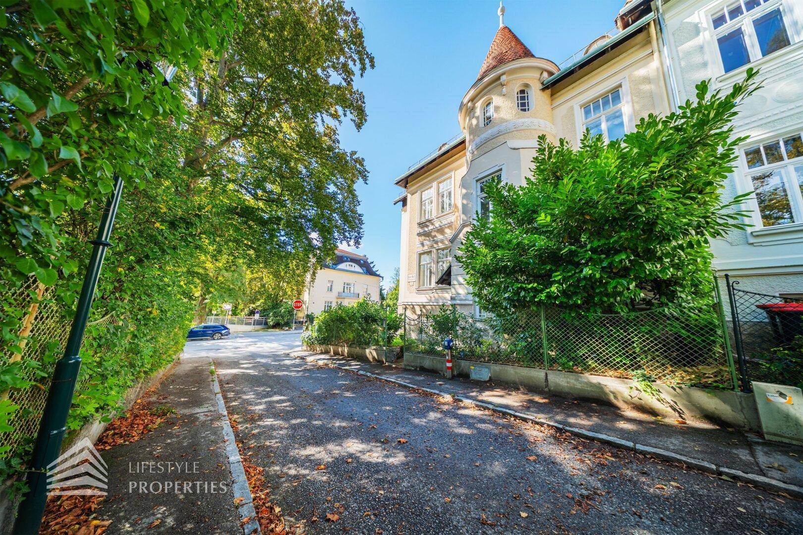 Seitenansicht der historischen Villa mit Turm und üppiger Bepflanzung entlang der Straße.