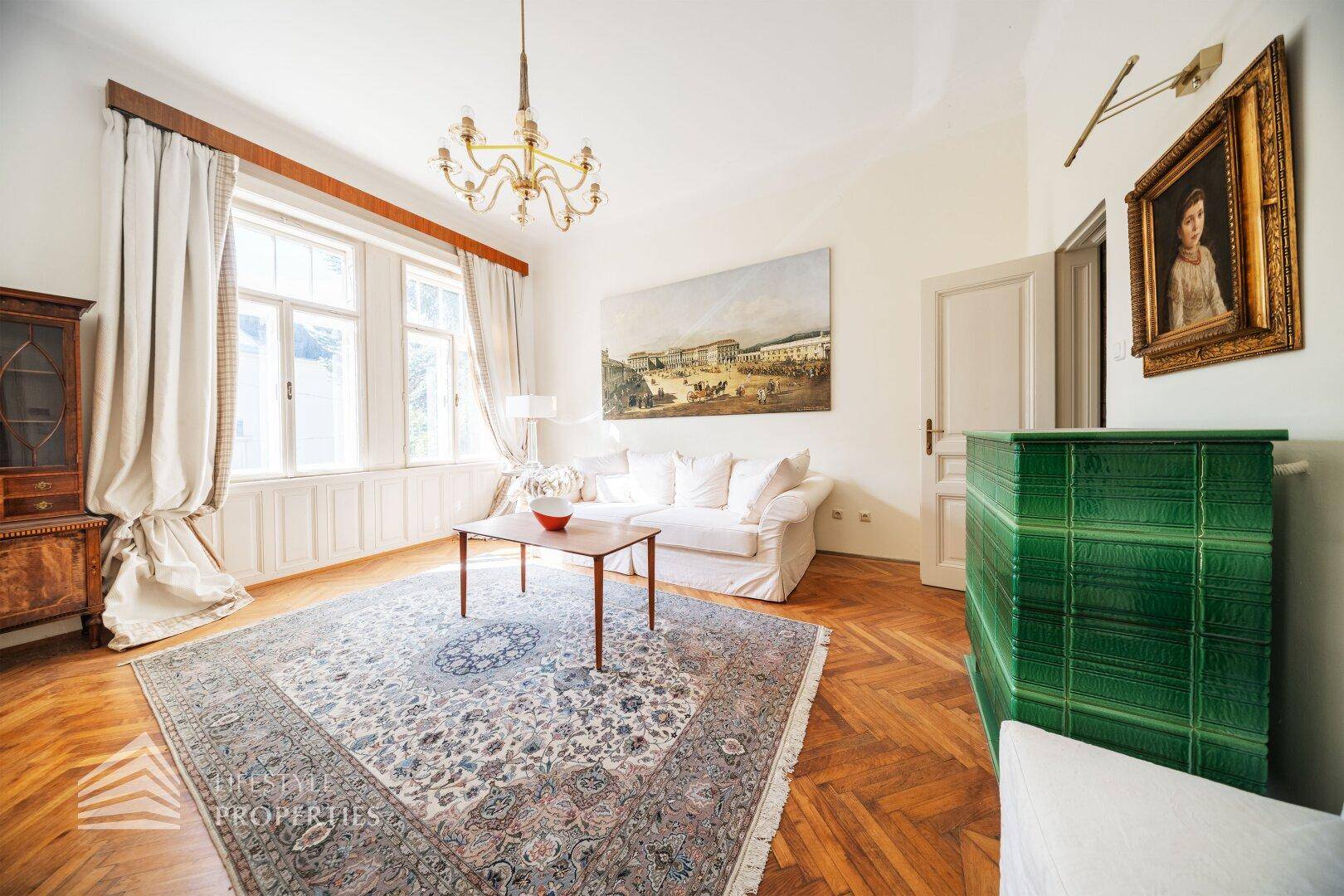 Elegantes Wohnzimmer mit Fischgrätparkett, Kachelofen und großen Fenstern mit Blick ins Grüne.