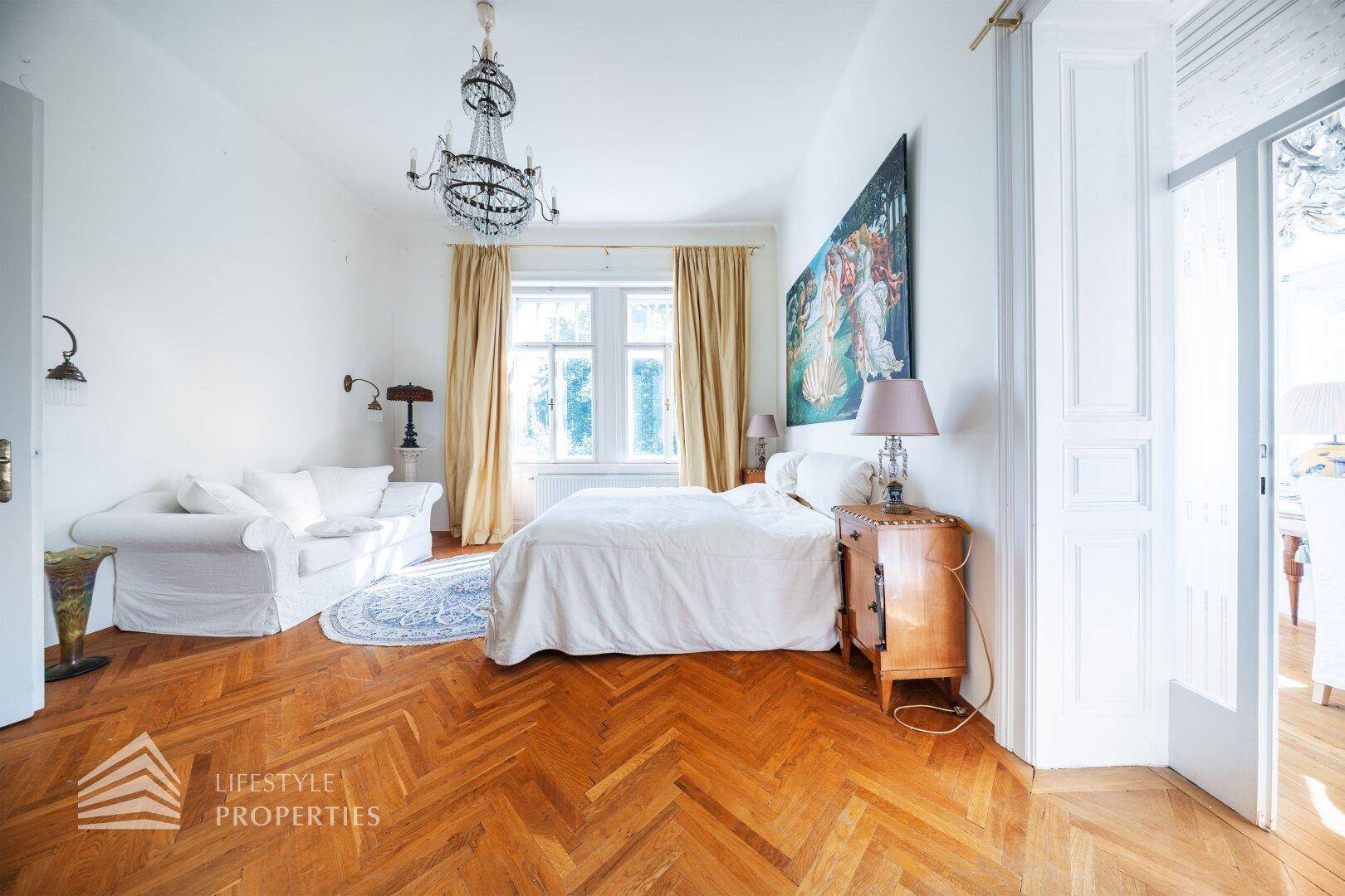 Geräumiges Schlafzimmer mit Fischgrätparkett, Kronleuchter und großem Bett.