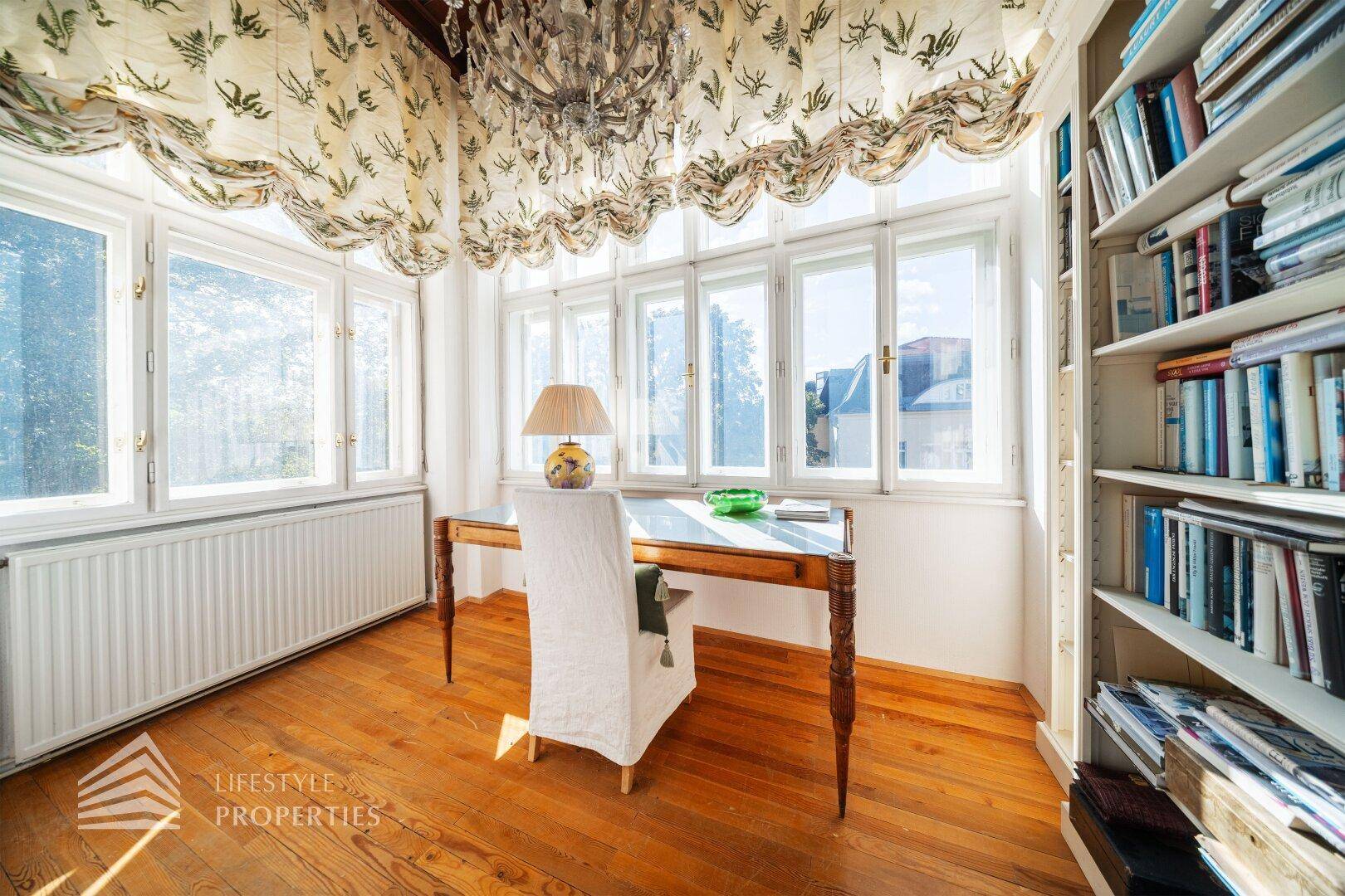 Helles Arbeitszimmer mit Holzschreibtisch, Bücherregal und vielen Fenstern.