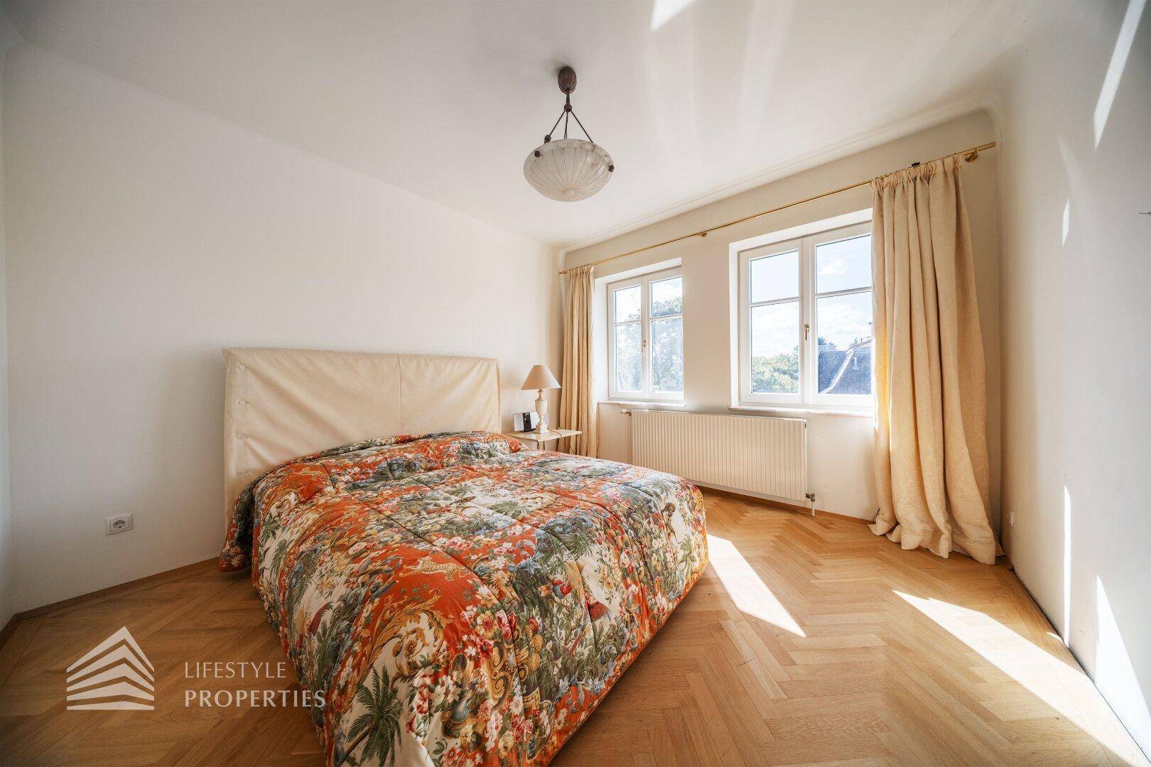 Helles Schlafzimmer mit Fischgrätparkett, großem Bett und Vorhängen.