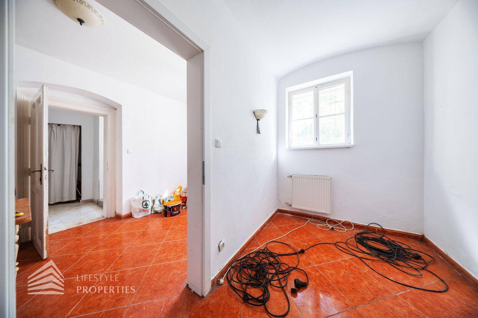 Kleiner Raum mit Terrakottafliesen und Fenster, derzeit als Abstellraum genutzt.