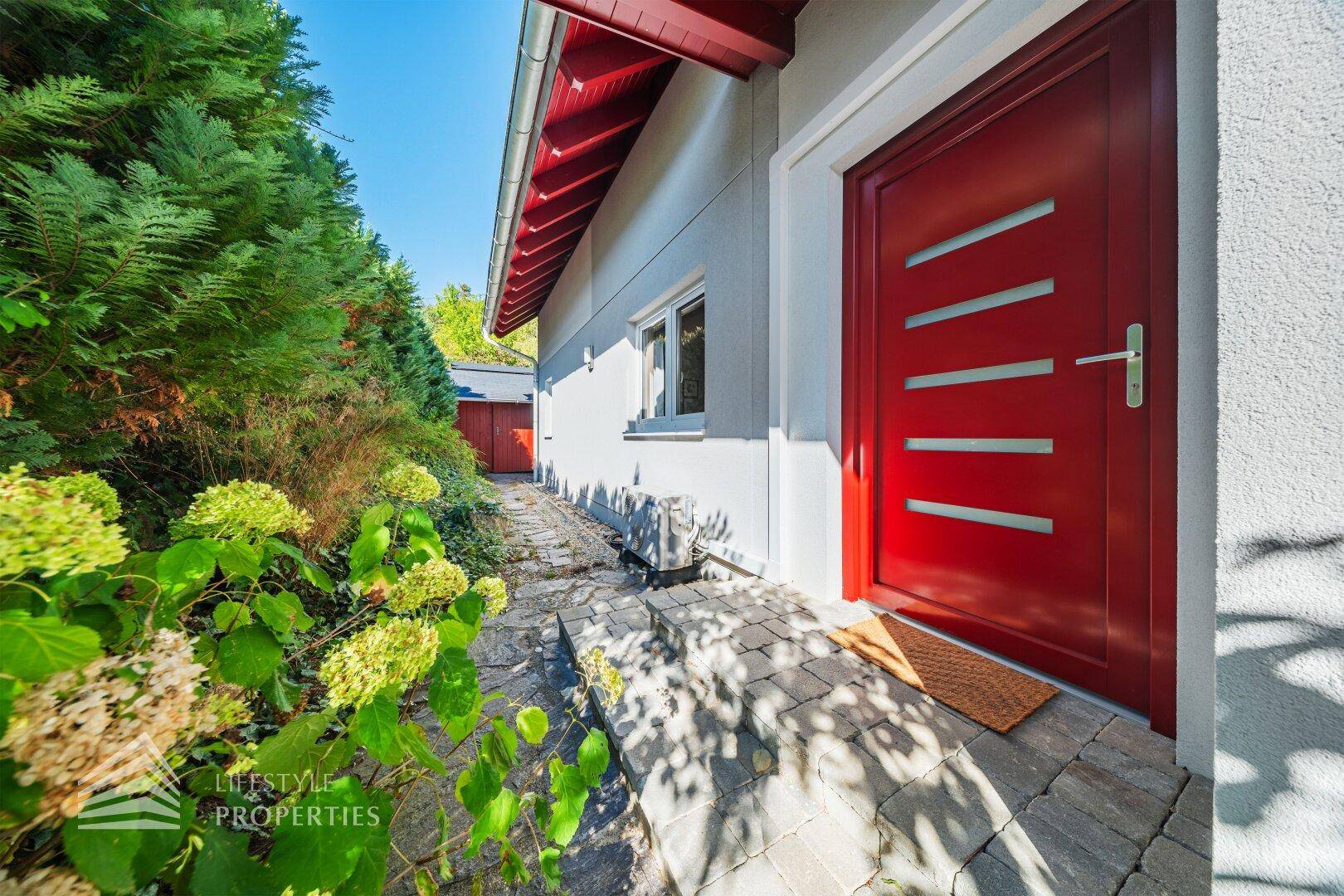 Rote Eingangstür mit modernen Glasstreifen und gepflastertem Zugangsbereich.
