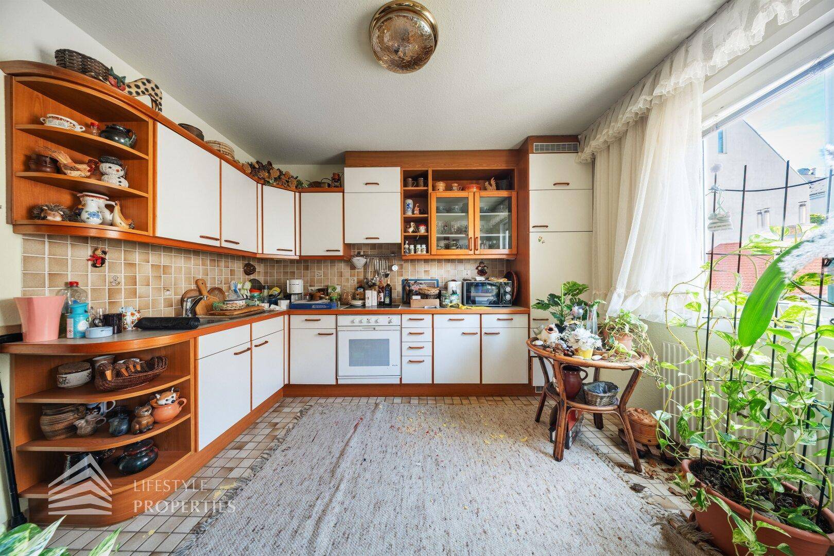 Funktionale Küche mit weißen Schränken, Fliesenboden und Pflanzen am Fenster.
