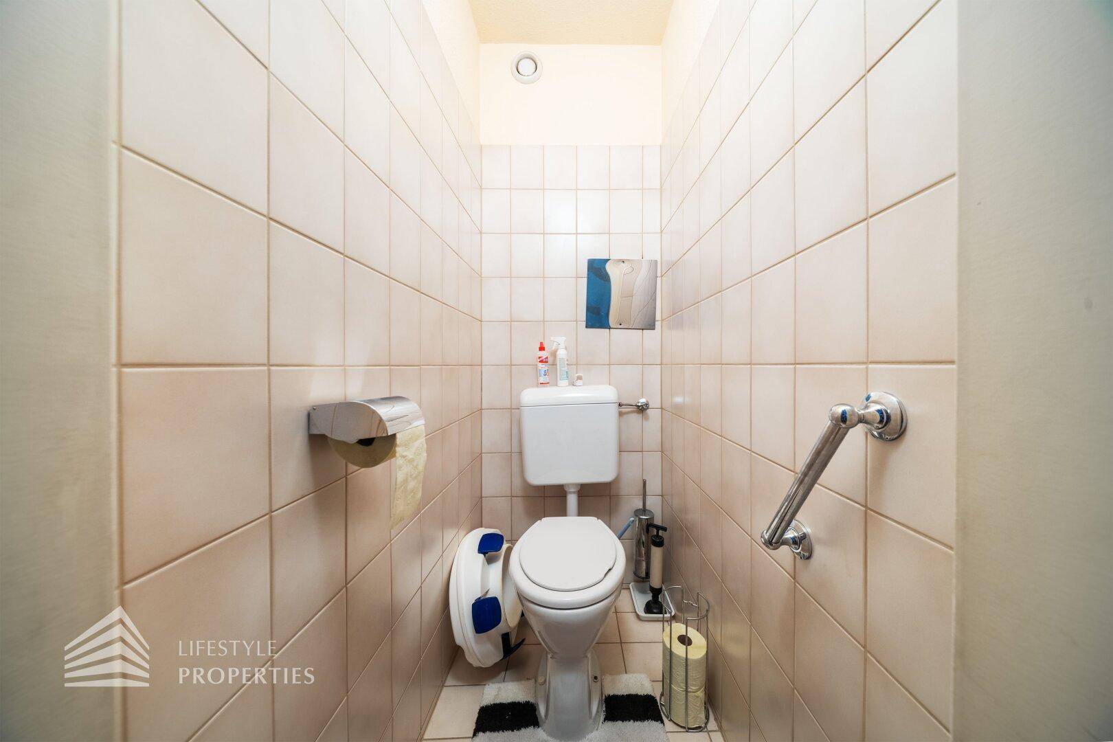 Separate Toilette mit Fliesen an Wänden und Boden sowie Haltegriffen.