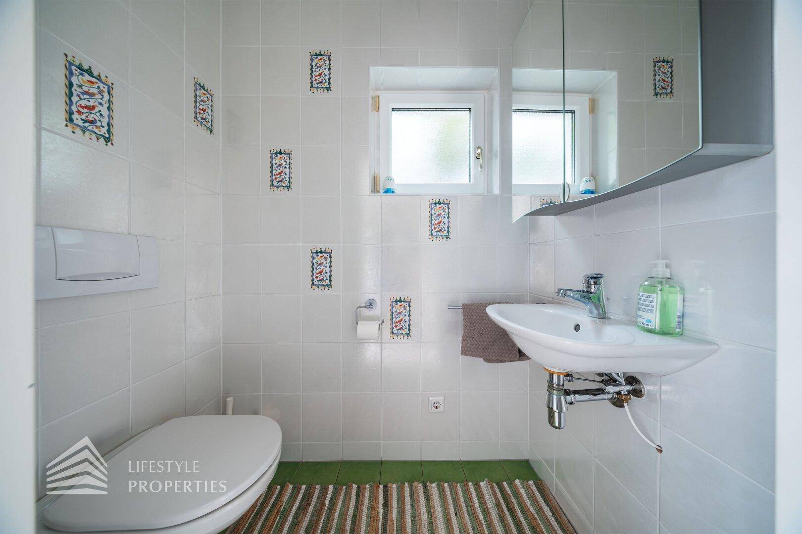 Gäste-WC mit weißen Fliesen, kleinem Waschbecken und Fenster für Belüftung.