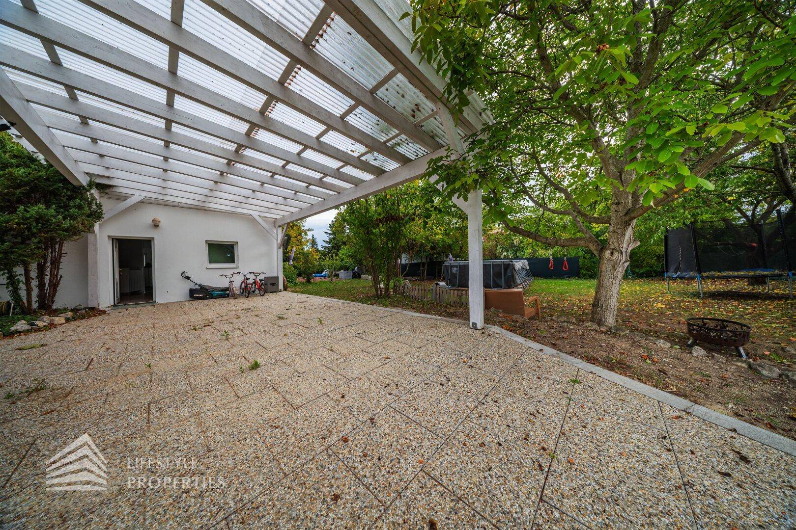 Großer Gartenbereich mit überdachter Terrasse, Rasenfläche und Trampolin.