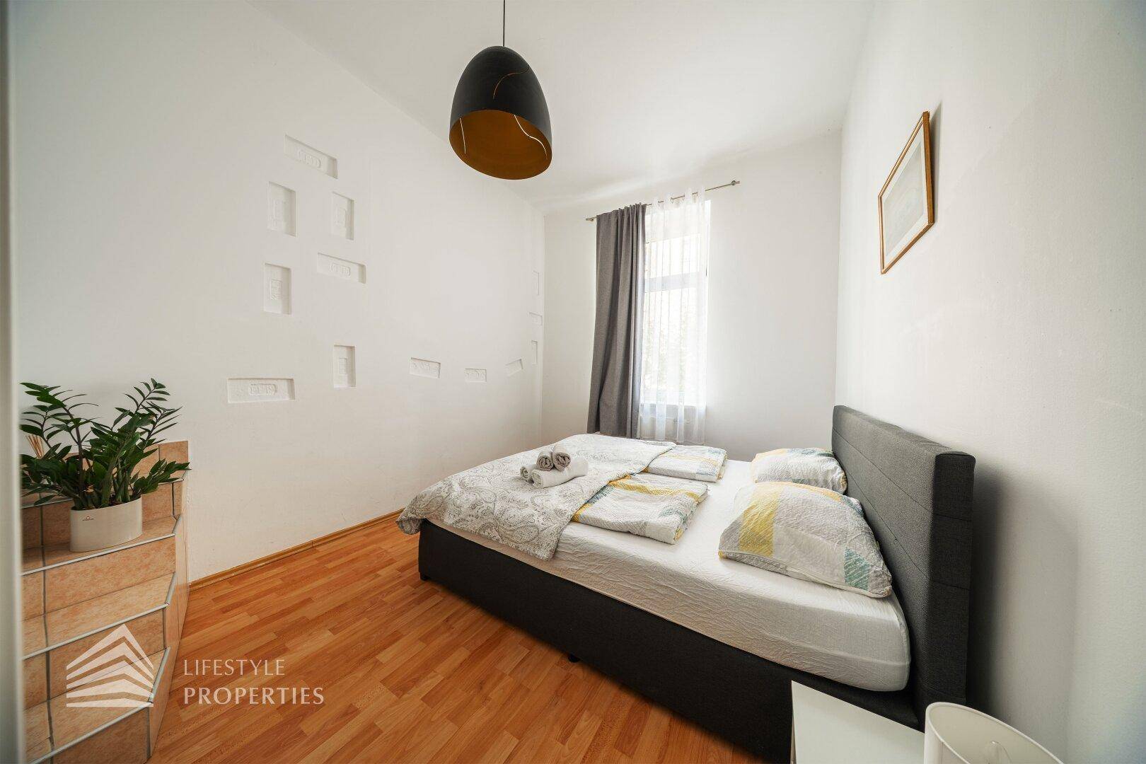 Helles Schlafzimmer mit Doppelbett, Holzboden und einem Fenster mit Vorhängen.