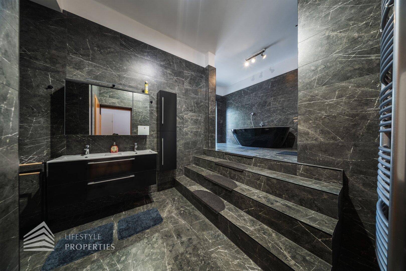 Modernes Badezimmer mit dunklen Marmorfliesen, Doppelwaschbecken und Stufen zur Badewanne.