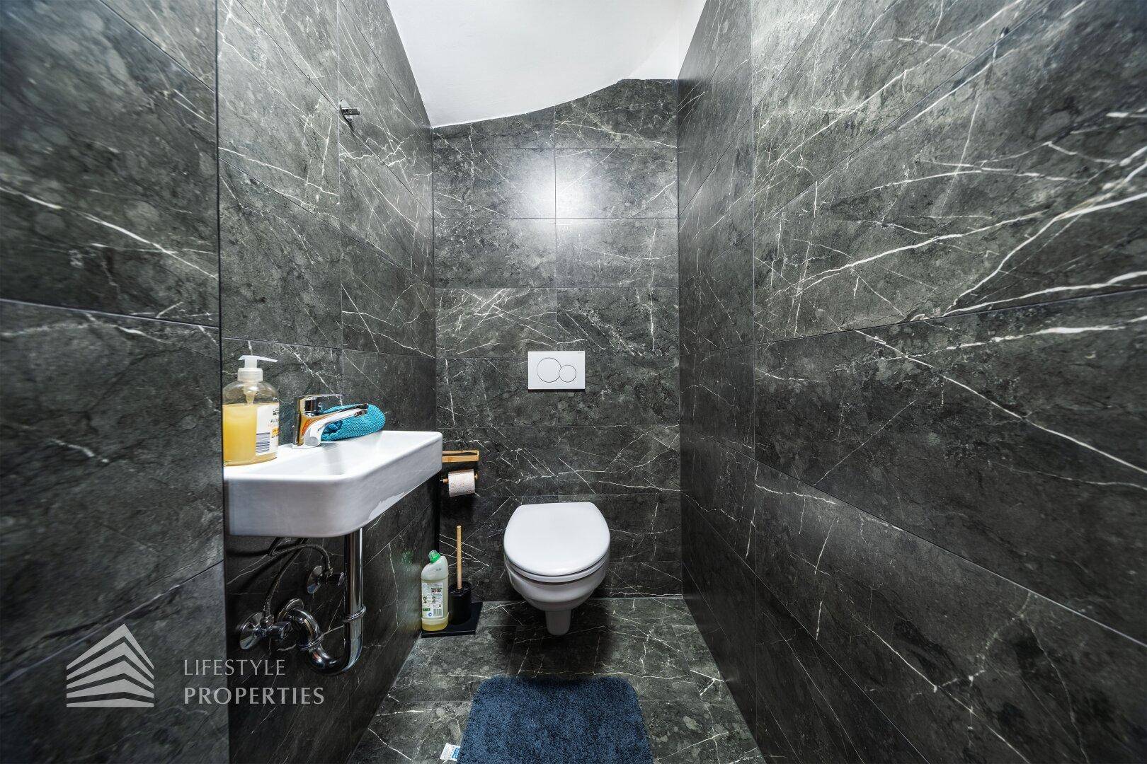 Modernes Gäste-WC mit dunklen Marmorfliesen, wandmontierter Toilette und kleinem Waschbecken.