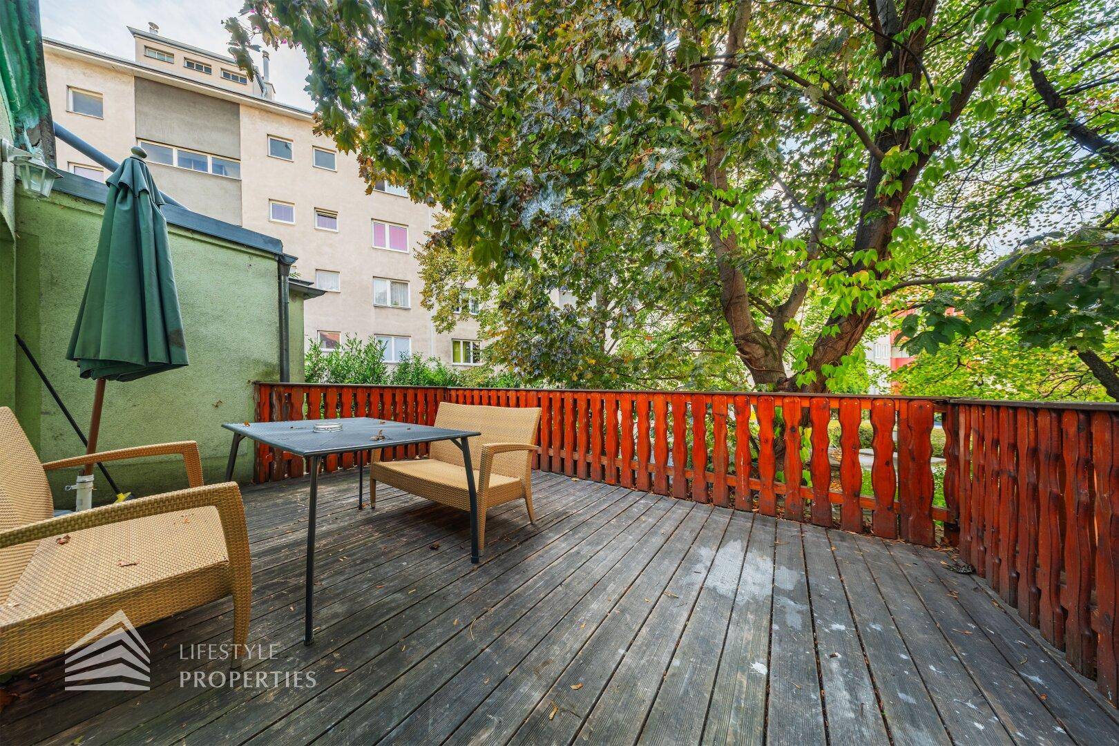 Holzterrasse mit Sitzgelegenheiten und Blick auf Bäume und umliegende Gebäude.