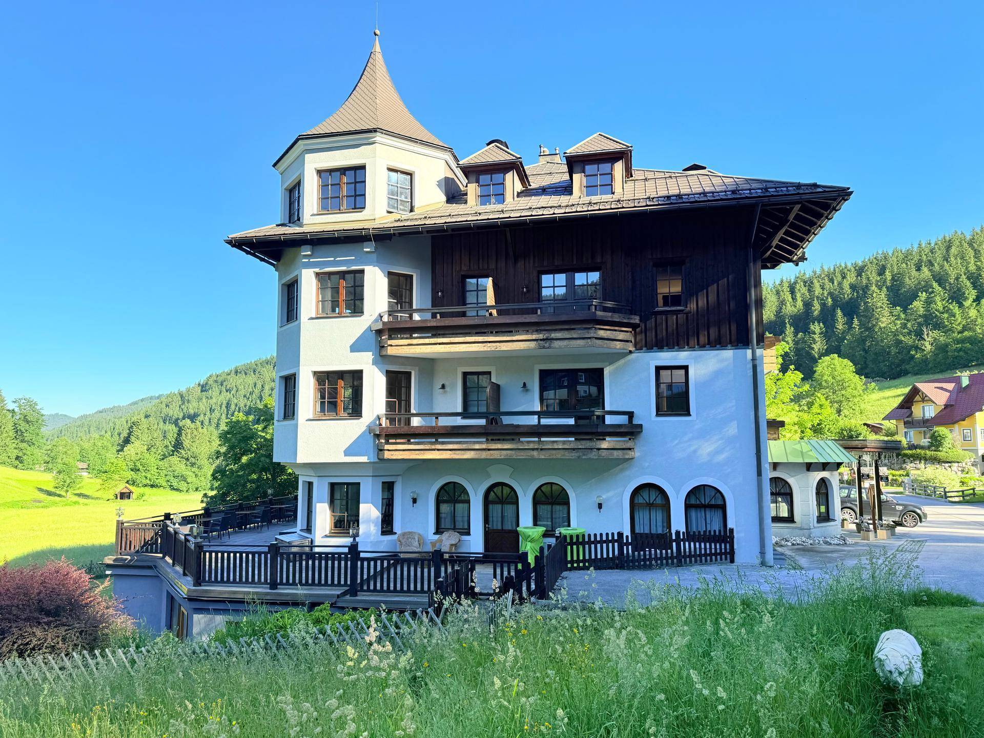Charmantes alpines Gebäude mit Turm und mehreren Balkonen, eingebettet in eine grüne Landschaft.