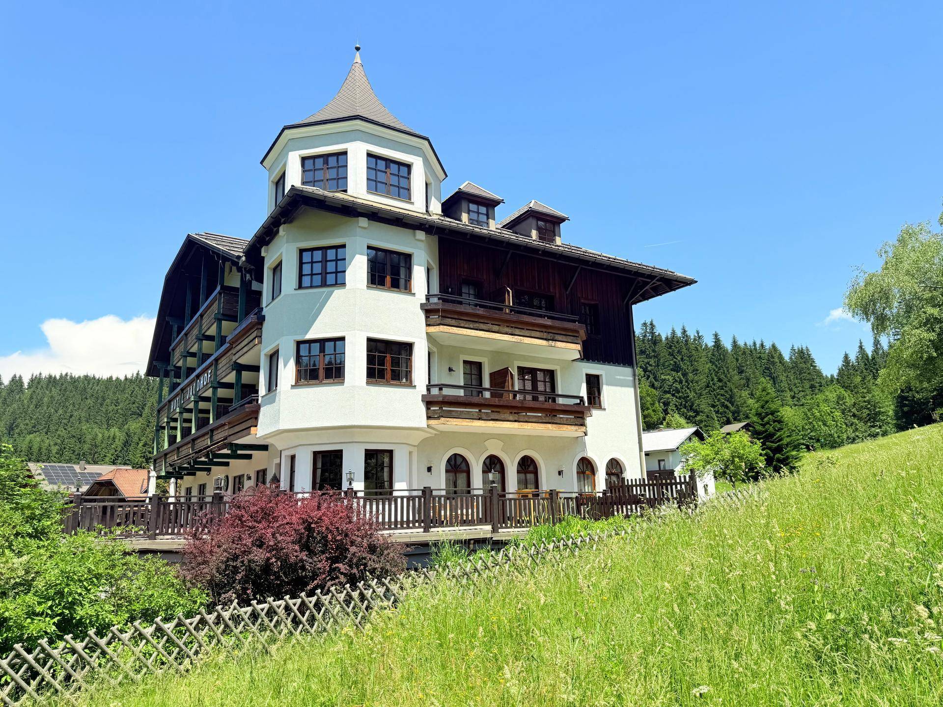 Traditionelles Gebäude im alpinen Stil mit Turm und Balkonen, umgeben von grünen Hügeln.
