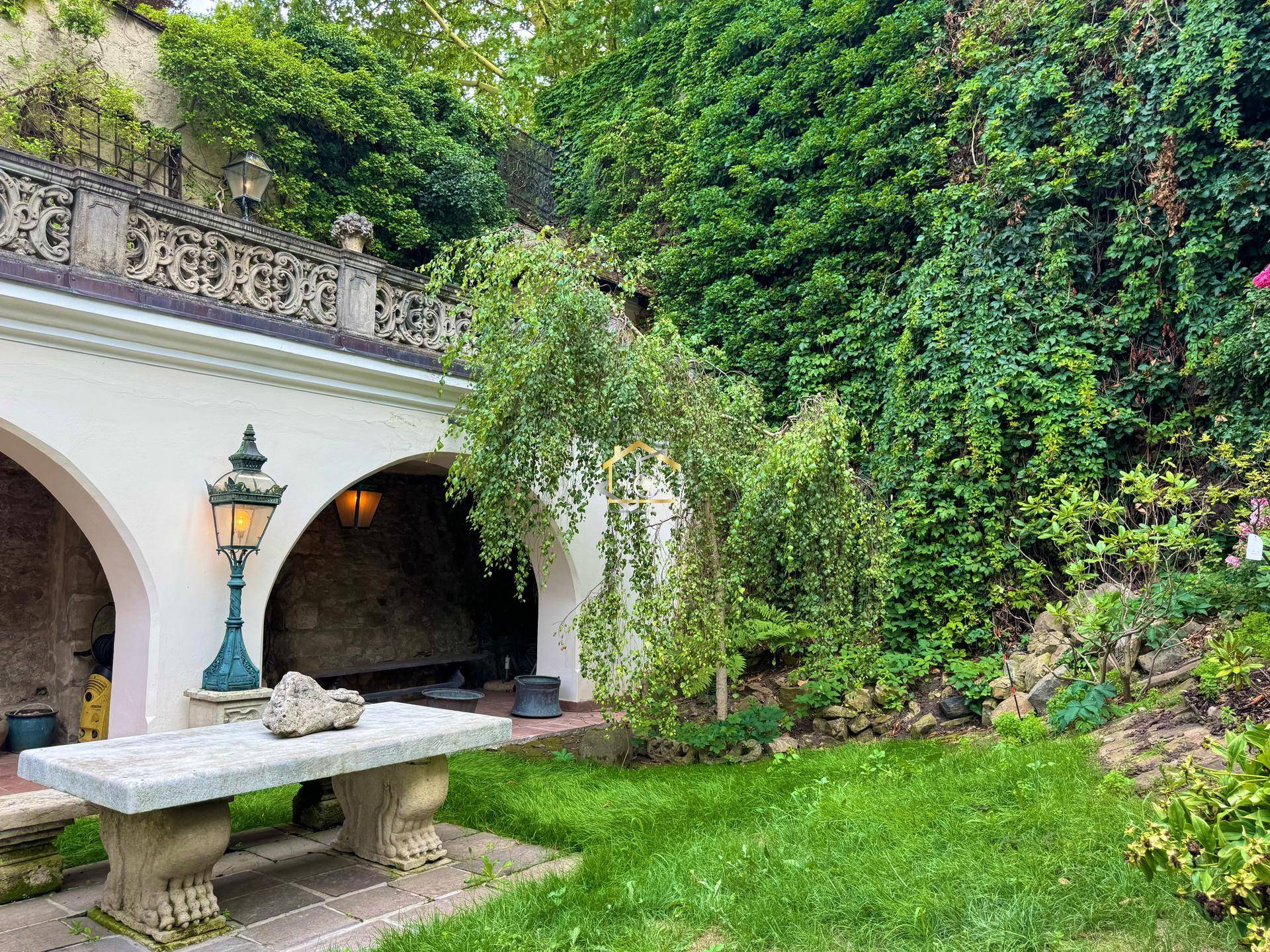 Idyllischer Gartenbereich mit Steinbank, üppigem Efeu an der Wand und historischen Bögen.