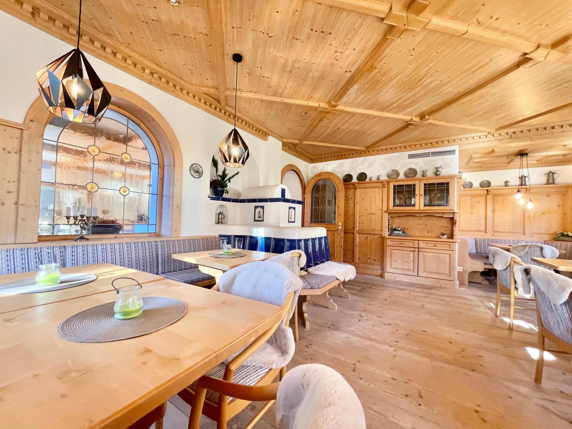 Traditioneller Speisesaal mit Holzboden, Holztischen und einem Kachelofen, beleuchtet durch große Fenster.