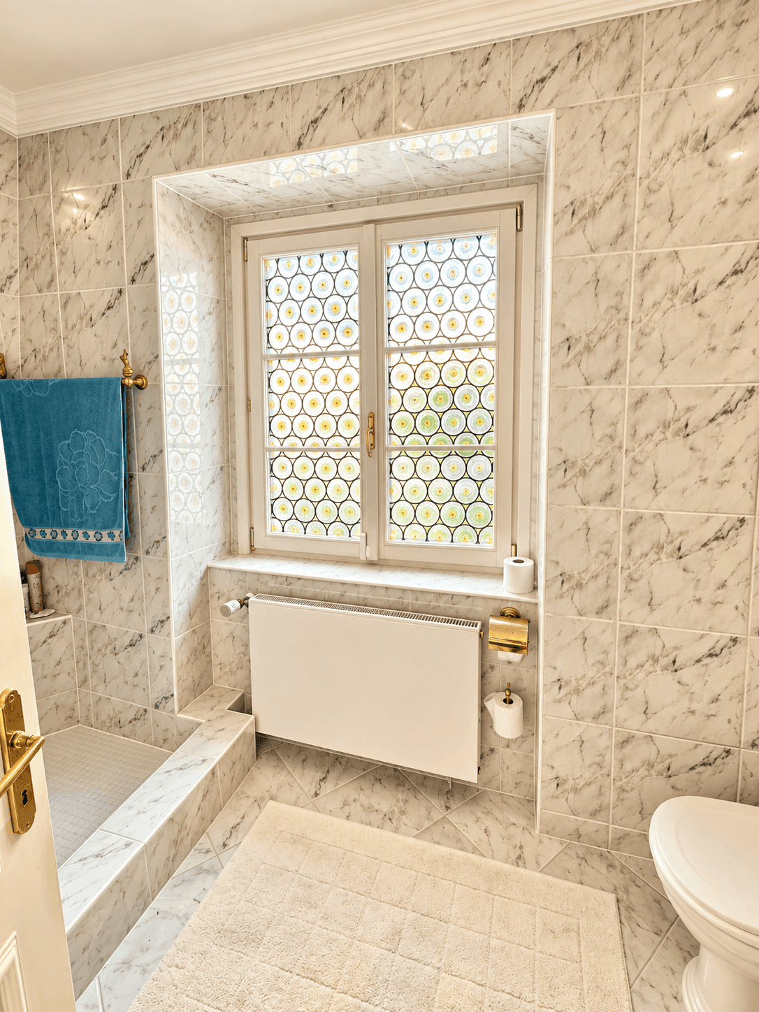 Helles Badezimmer mit Marmorfliesen, Fenster und einer Toilette, ausgestattet mit Heizkörper.
