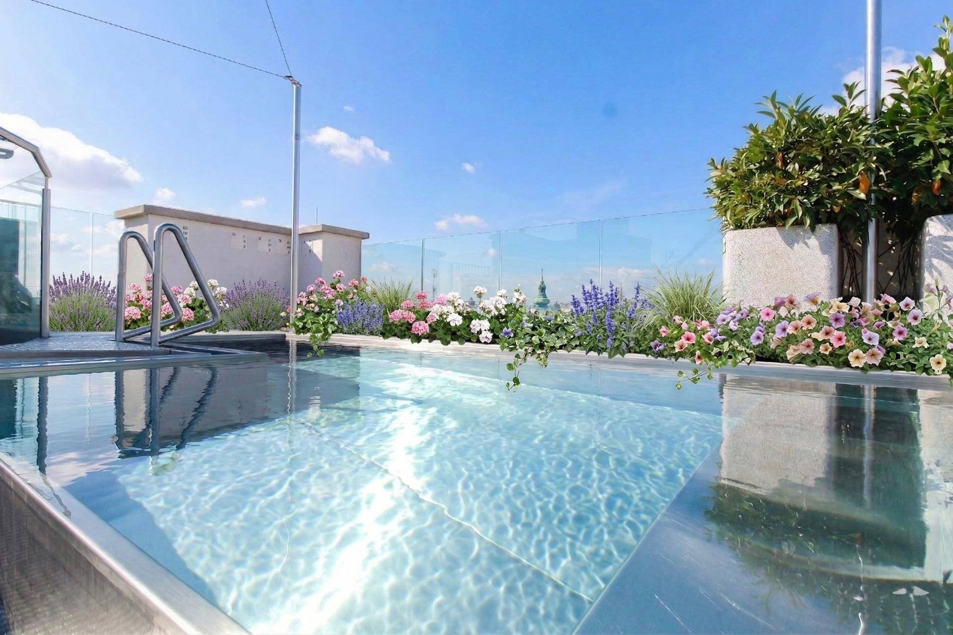 Dachterrasse mit Infinity-Pool, Blumenbeeten und weitem Stadtblick unter blauem Himmel.