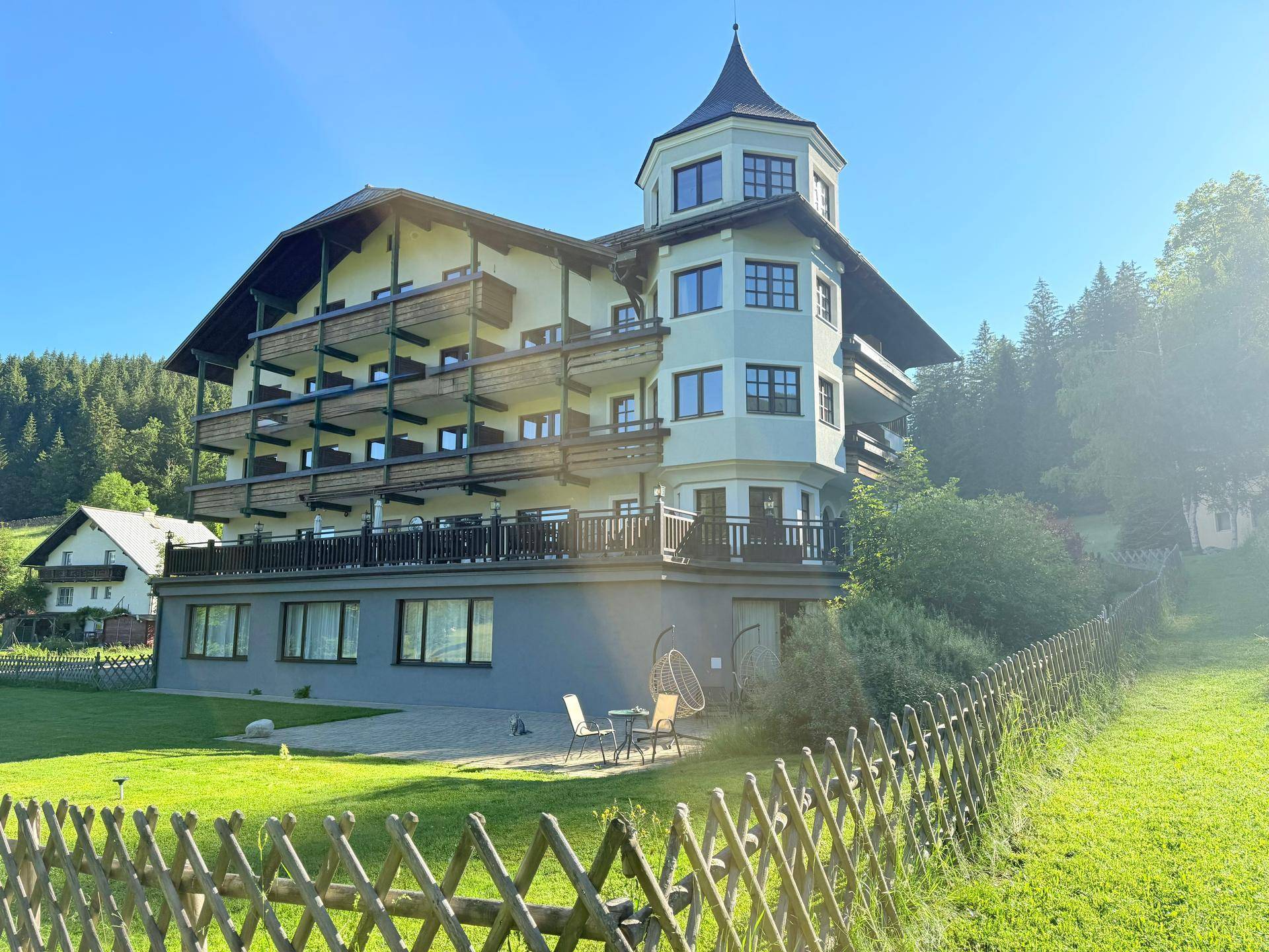 Alpines Gebäude mit Turm und Balkonen, umgeben von einer grünen Wiese unter blauem Himmel.