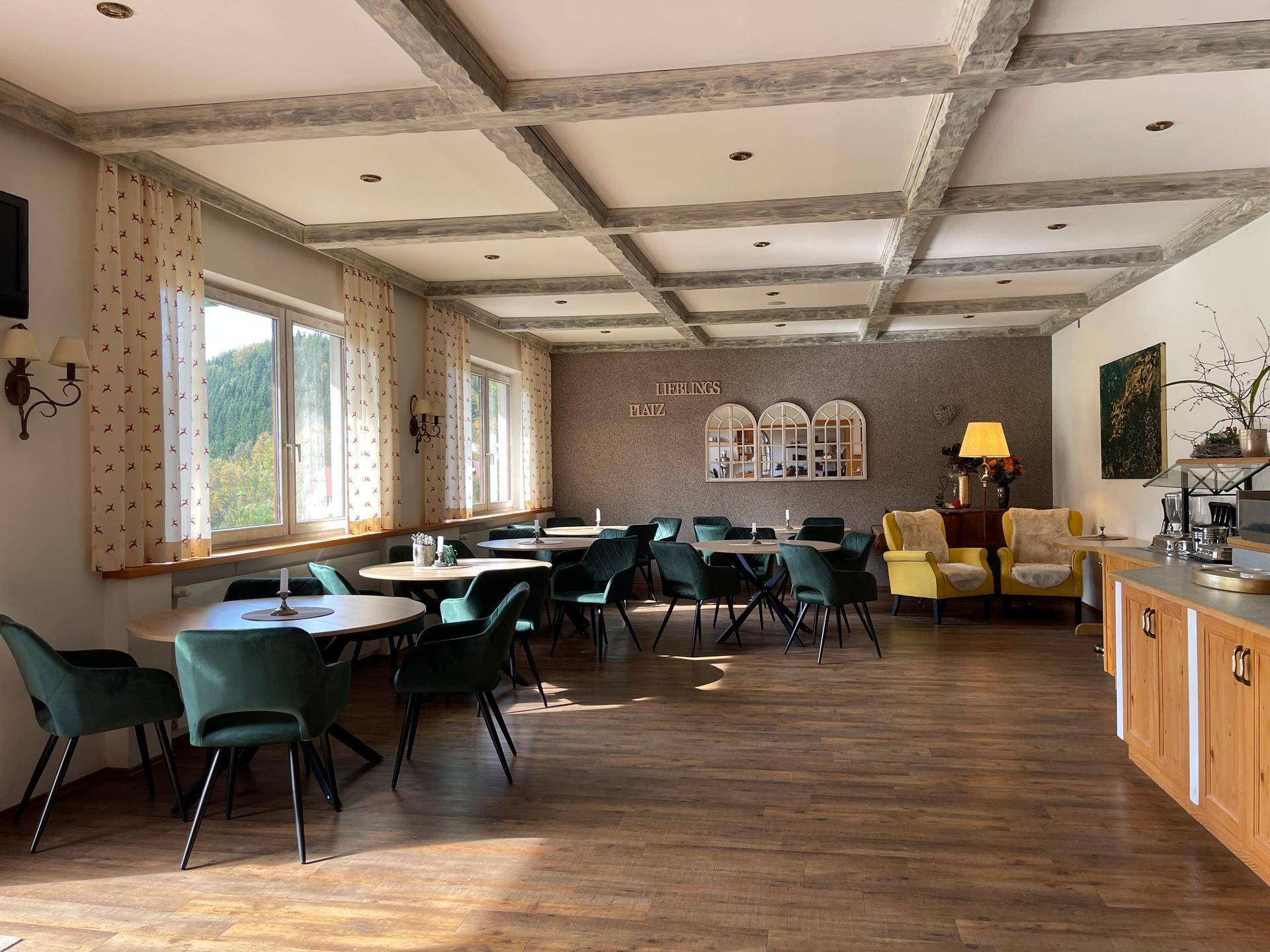 Heller Speisesaal mit großen Fenstern, Holzboden und grünen Samtstühlen.