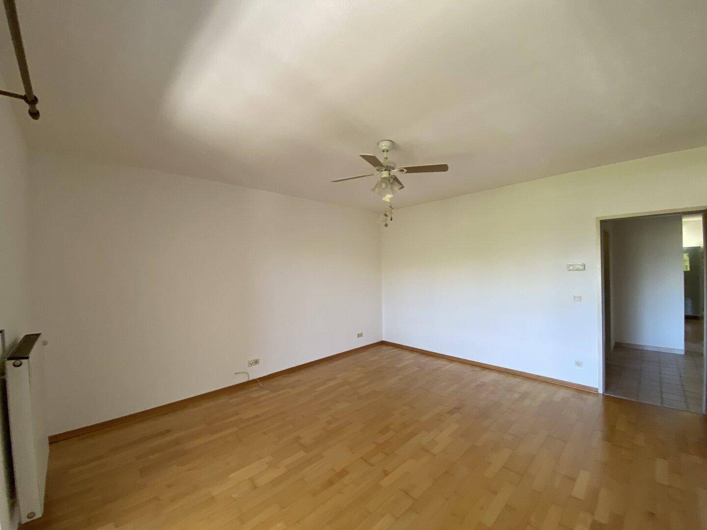 Geräumiges, leeres Wohnzimmer mit hellem Parkettboden und Deckenventilator, gut beleuchtet.