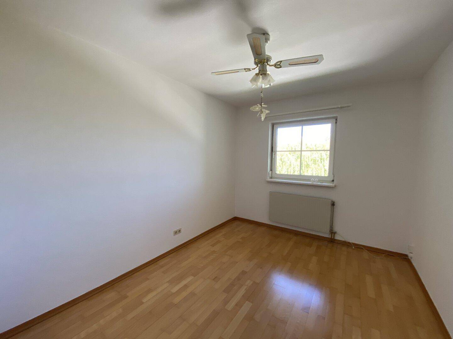 Helles Schlafzimmer mit Parkettboden, Fenster und einem Deckenventilator, gut gepflegt.