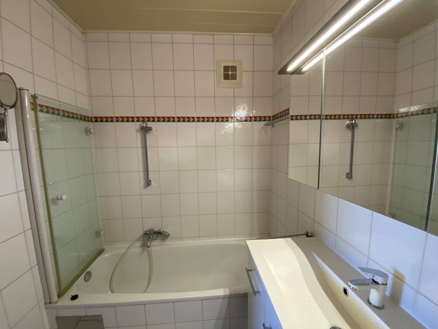 Gefliestes Badezimmer mit Badewanne, Duschwand, Einzelwaschbecken und Spiegelschrank, Standardausstattung.