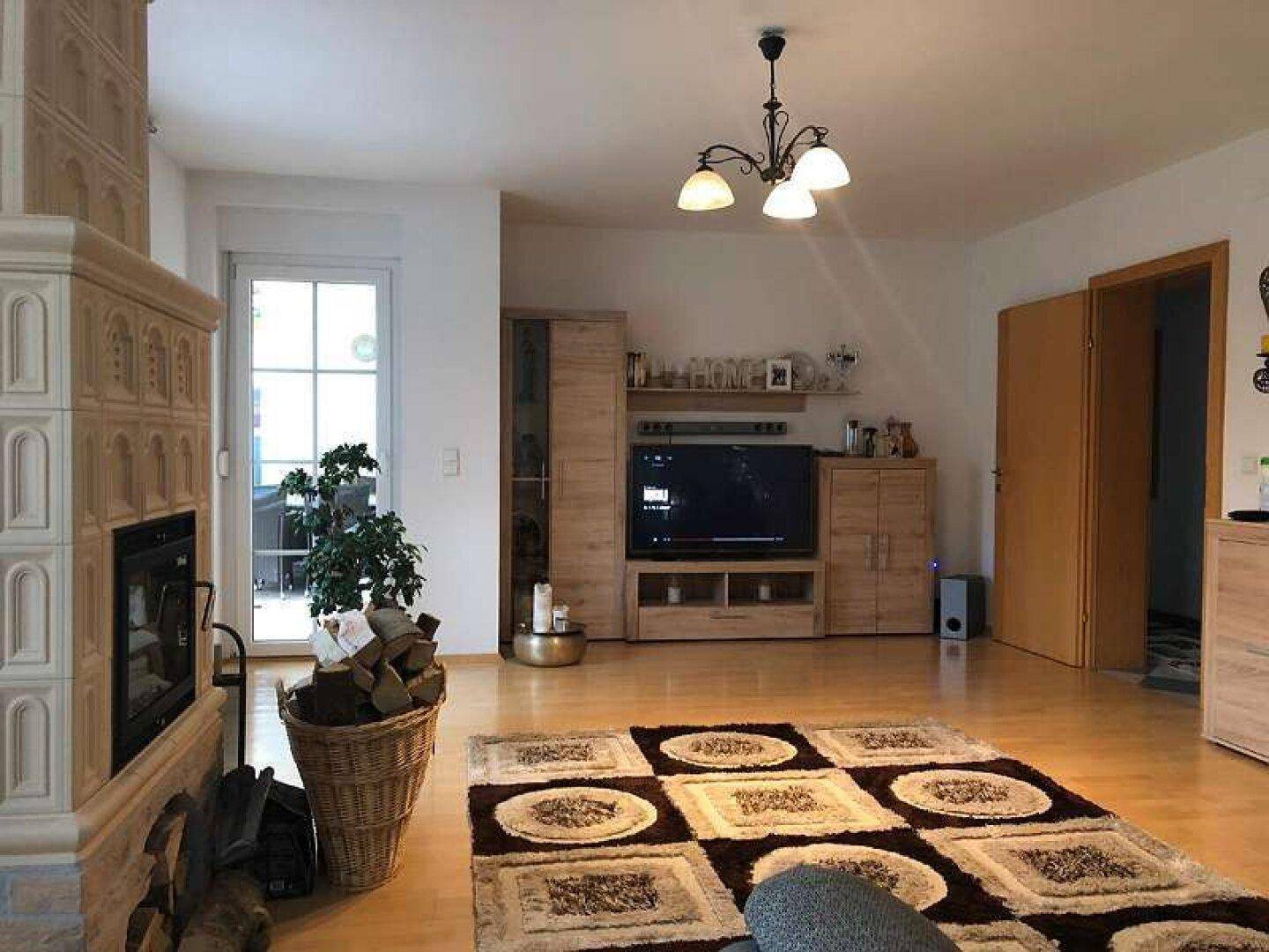 Wohnzimmer mit Kachelofen, Holzboden und Zugang zur Terrasse, eingerichtet mit Holzmöbeln.