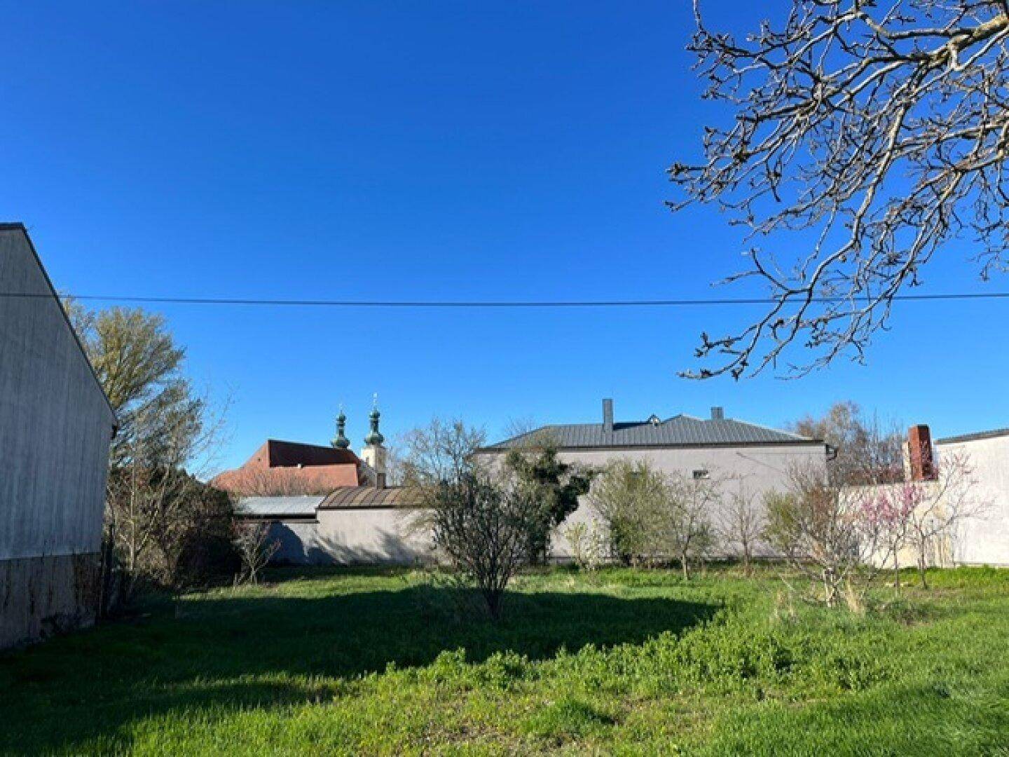 Grüne Gartenfläche mit Bäumen, umgeben von Gebäuden und einem Kirchturm am Horizont.