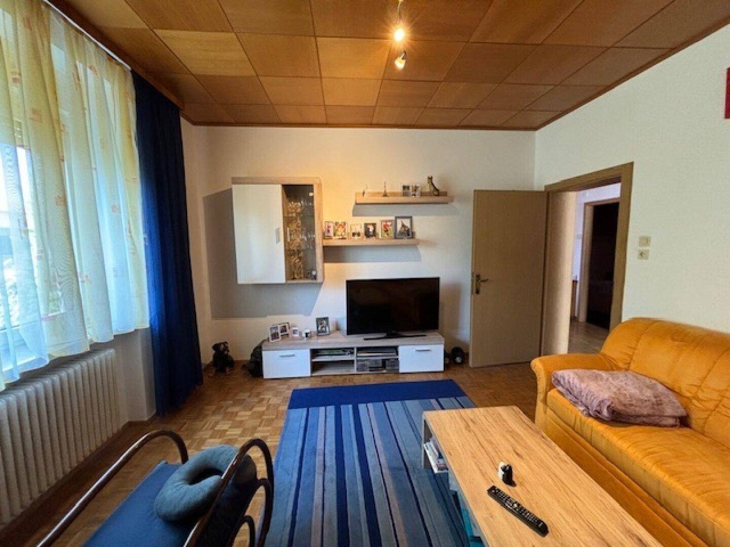 Wohnzimmer mit Fernseher, Holzdecke und Parkettboden, hell durch große Fenster.