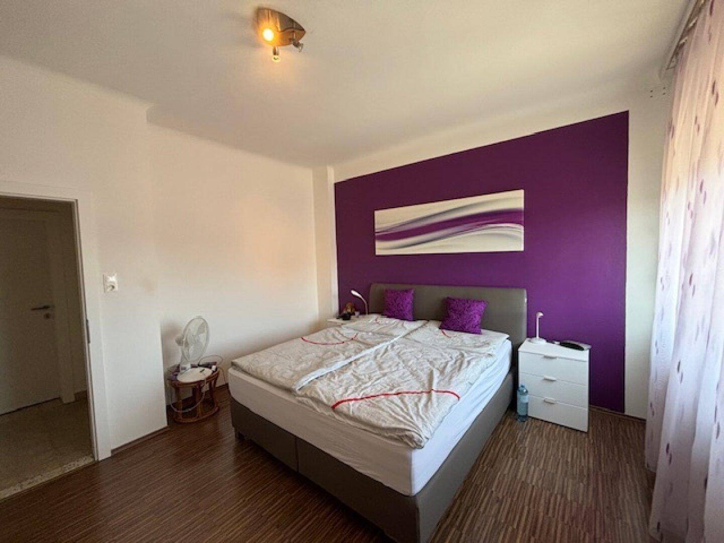 Schlafzimmer mit lila Akzentwand, Doppelbett und modernem Kunstwerk über dem Bett.