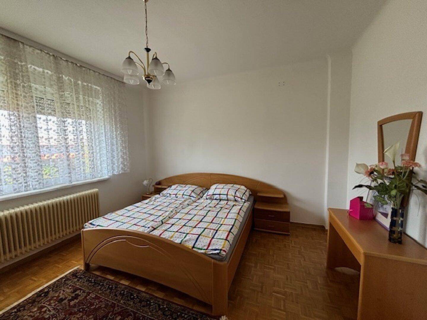 Helles Schlafzimmer mit Doppelbett, Holzmöbeln und großem Fenster mit Gardinen.