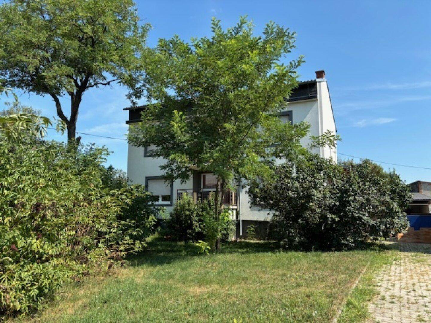 Wohnhaus mit viel Grün und Bäumen im Vorgarten unter strahlend blauem Himmel.