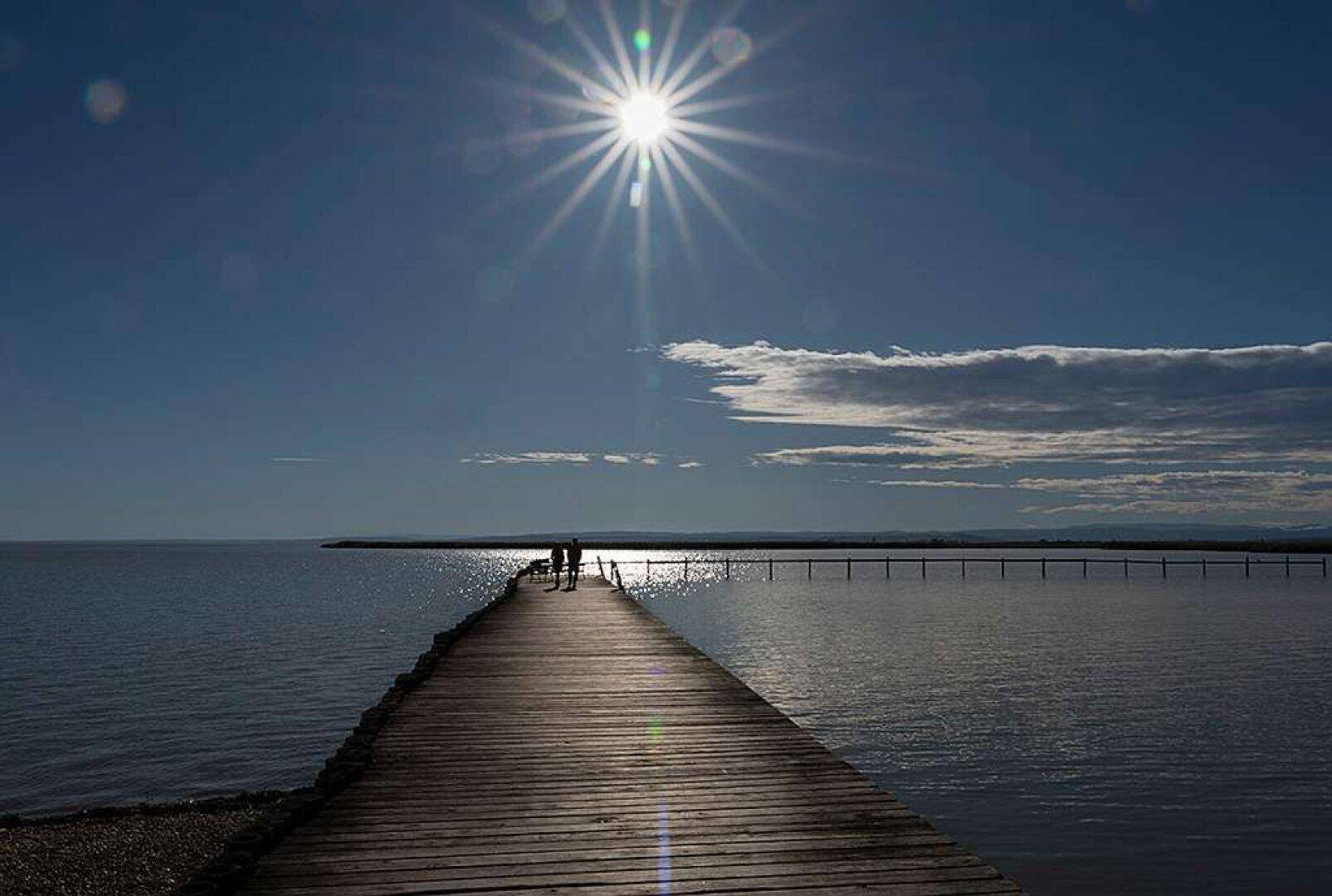 Langer Holzsteg führt in einen weiten See unter strahlendem Sonnenschein und blauem Himmel.